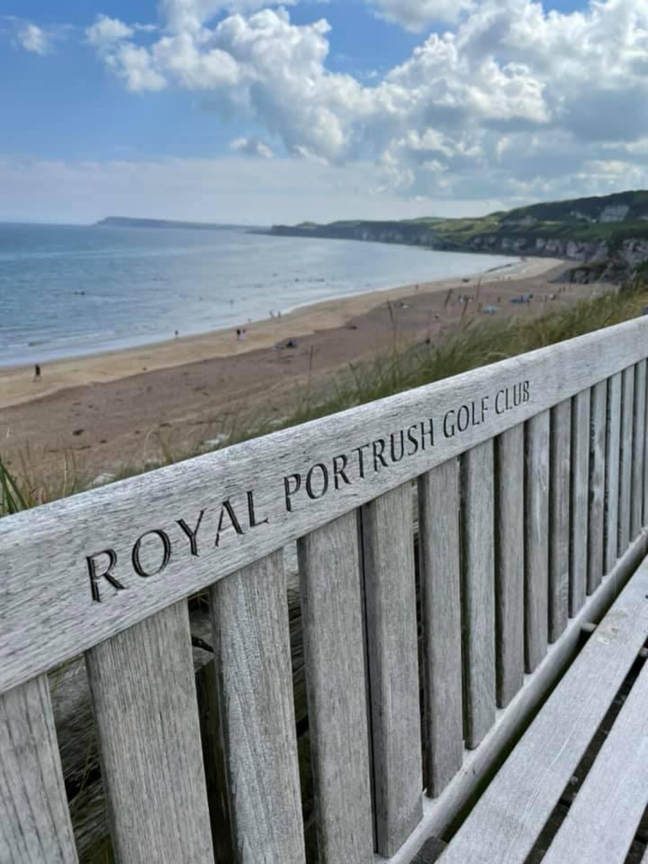 Wooden bench engraved Royal Portrush Golf Club overlooking a beach and cliffs
