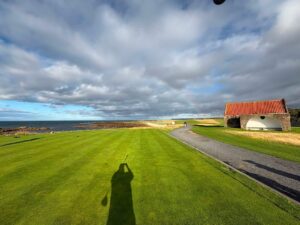A coastal links tee in Scotland beside a small stone shelter with a red-tiled roof, the sea beyond