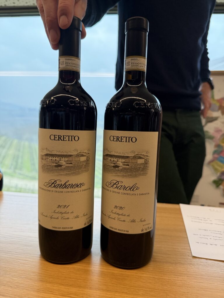 Two bottles of Ceretto wine — a Barbaresco 2021 and a Barolo 2020 — held up at a tasting in Piedmont, Italy, vineyards behind